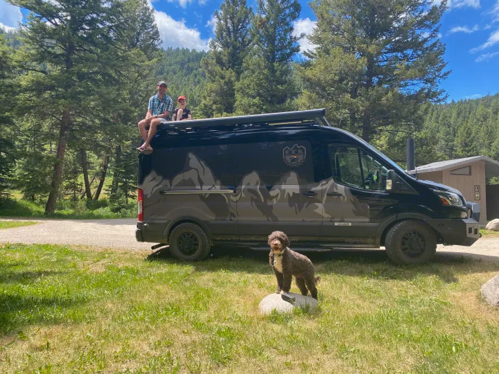 Family enjoying their black RV rental. Dad & son on the roof, dog on a rock, surrounded by green forest & mountains.. Ford Custom Transit 2020