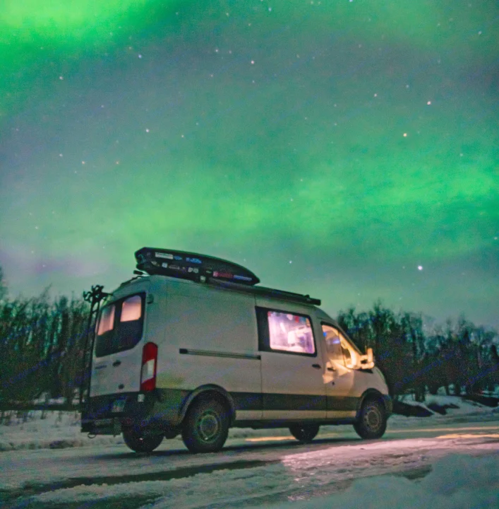 An rv rental glows on a snowy road, its interior lit, set against the stunning green Northern Lights and a starry night sky.. Ford Transit Custom 2015