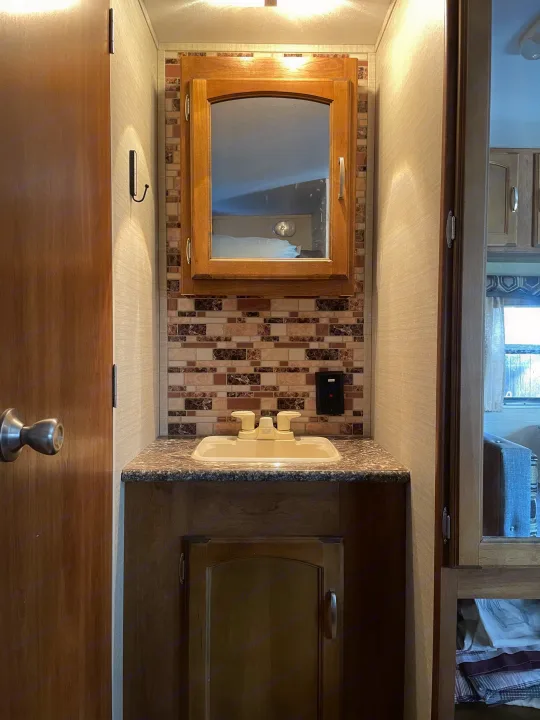 Inside an RV rental, this bright bathroom features a compact vanity with a sink, wooden mirror cabinet, and tiled backsplash.. Keystone RV Springdale 2016