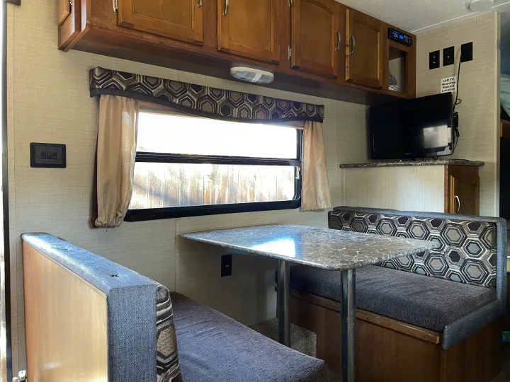 Interior of an rv rental: a well-lit dinette area with patterned benches, table, overhead cabinets, and a mounted TV.. Keystone RV Springdale 2016