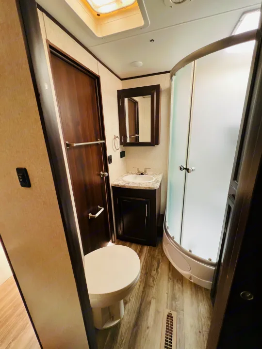 RV rental bathroom: A compact space with a toilet, sink, mirror, and curved frosted glass shower stall. Wood-look floor.. Jayco Jay Flight 2019