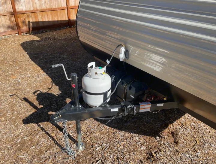 Front view of an RV rental, showing its hitch with a white propane tank, black battery boxes, and jack on a wood chip surface.. Dutchmen Aspen Trail 2021