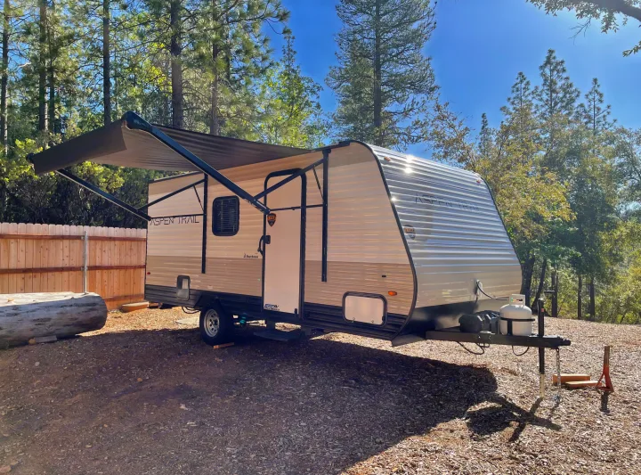 Aspen Trail travel trailer, ideal for an RV rental, parked outdoors with its awning extended at a rustic campsite among tall trees.. Dutchmen Aspen Trail 2021