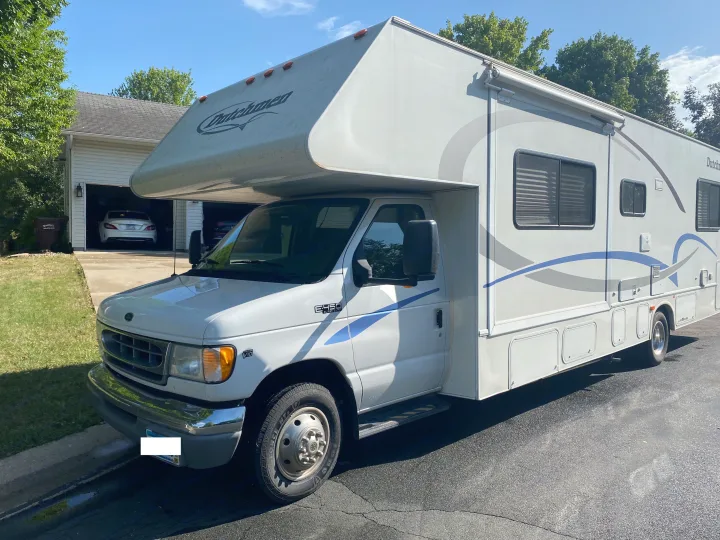 This white Class C RV rental, a Ford E-450 Dutchmen motorhome, is parked on asphalt with a garage and trees in the background.. Dutchmen Four Winds 2003