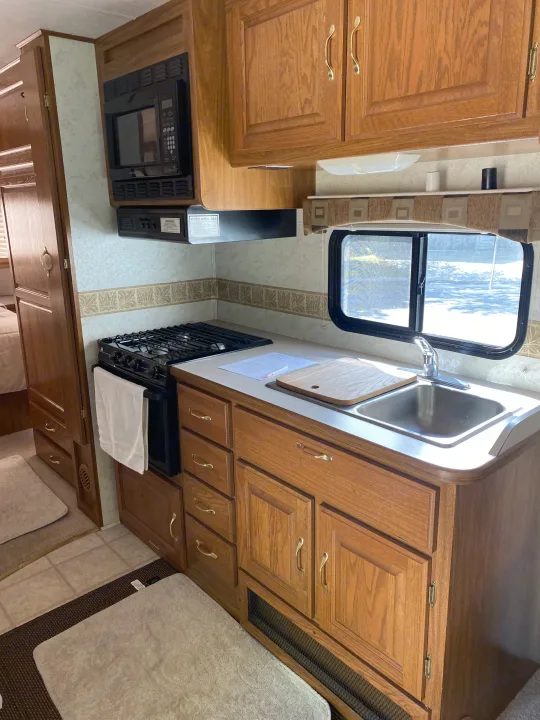 Bright rv rental kitchen featuring a microwave, gas stove, sink, wooden cabinets, and a sunlit window.. Dutchmen Four Winds 2003