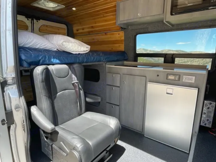 Modern RV rental interior: upper bunk bed, black seat, sleek kitchenette with fridge, & stunning mountain views from the window.. RAM Promaster ALPINE X Edition OA15 2021