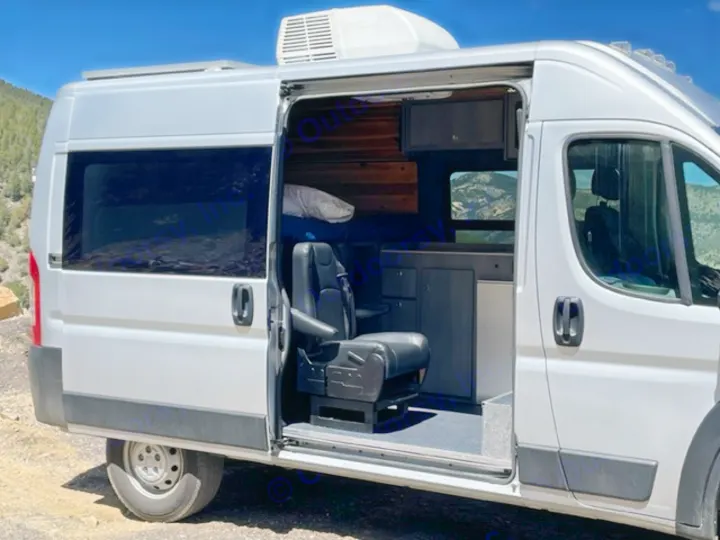 This silver camper van RV rental, with its side door open, reveals a swivel seat, kitchenette, and bed area, offering mountain views.. RAM Promaster ALPINE X Edition OA15 2021