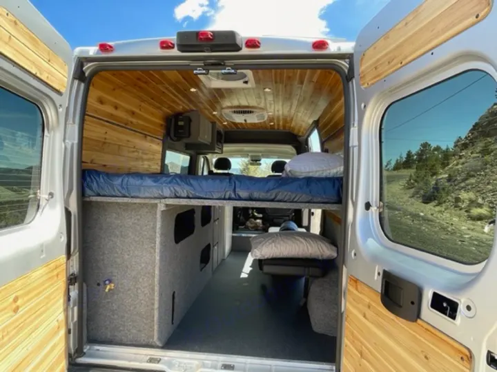 Open rear doors of an RV rental revealing a cozy, elevated bed, wooden ceiling, and carpeted storage units inside. Ready for travel!. RAM Promaster ALPINE X Edition OA15 2021