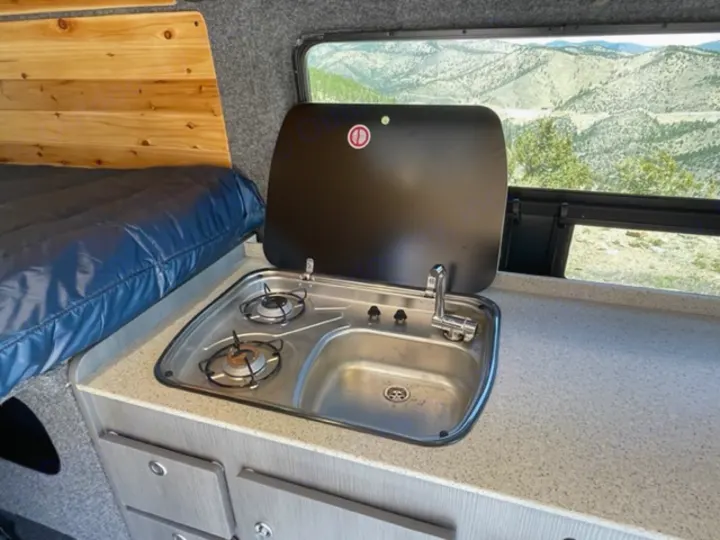 This RV rental features a compact kitchen with a two-burner stove, sink, nearby bed, and window with a scenic mountain view.. RAM Promaster ALPINE X Edition OA15 2021