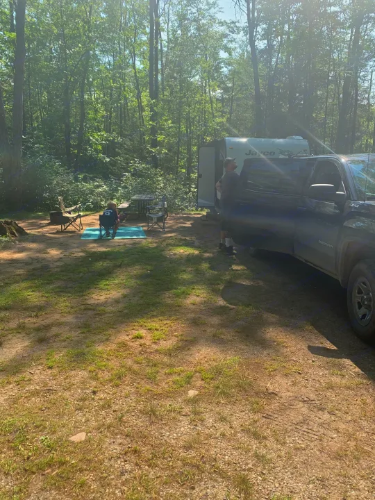 Sunny forest campsite with an RV rental and truck; a person sits by a picnic table on a blue mat.. Jayco Jay Flight 2020