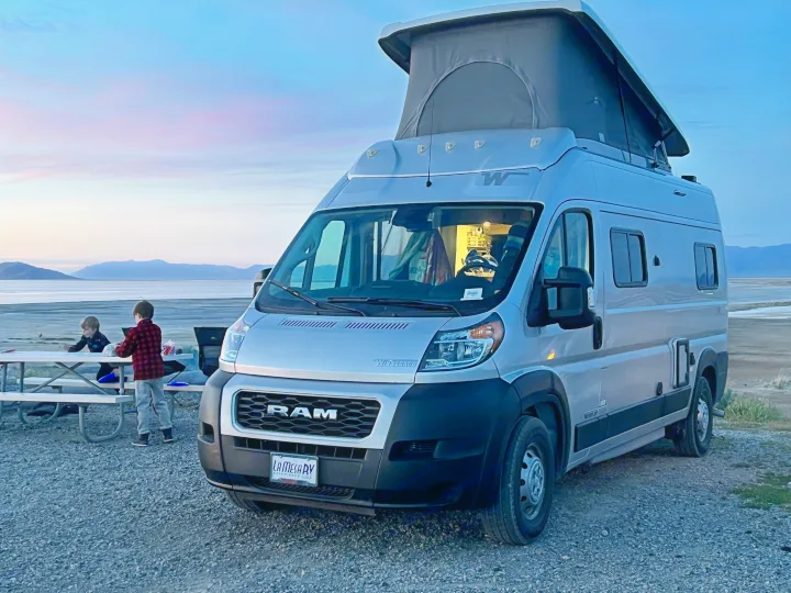 An RV rental camper van with a pop-top is parked by a lake at twilight, two children are at a picnic table nearby.. Winnebago Solis 2022