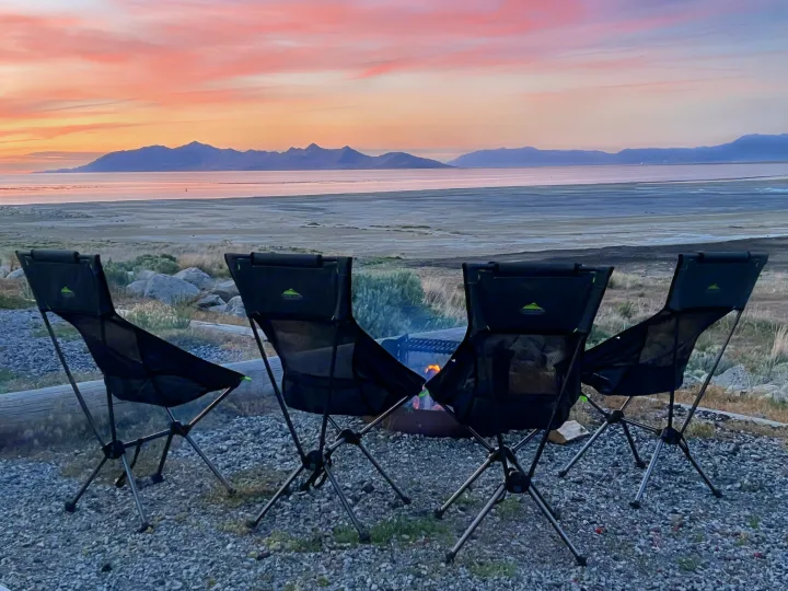 Four camp chairs by a fire pit, facing a beautiful sunset over distant mountains and a calm lake. Perfect RV rental site.. Winnebago Solis 2022