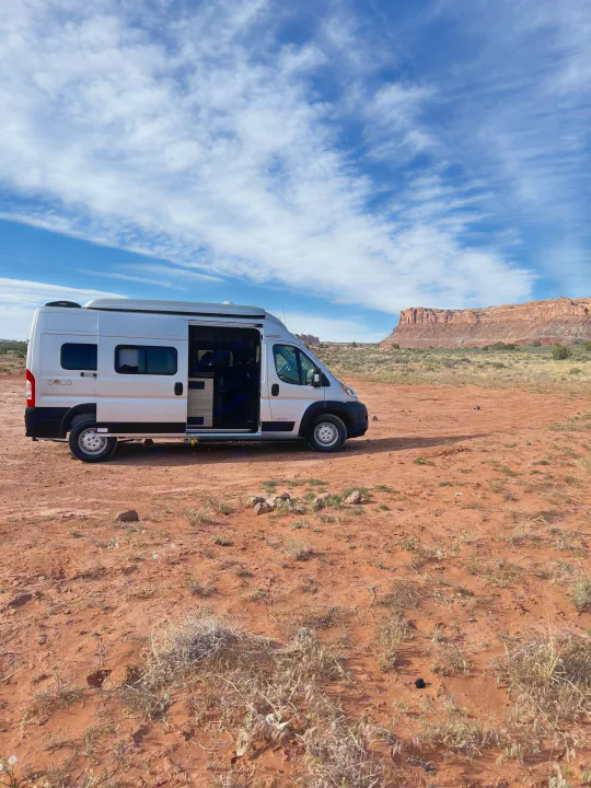 A white Winnebago Solis rv rental parked on red desert earth with an open side door, facing a majestic mesa under blue skies.. Winnebago Solis 2022