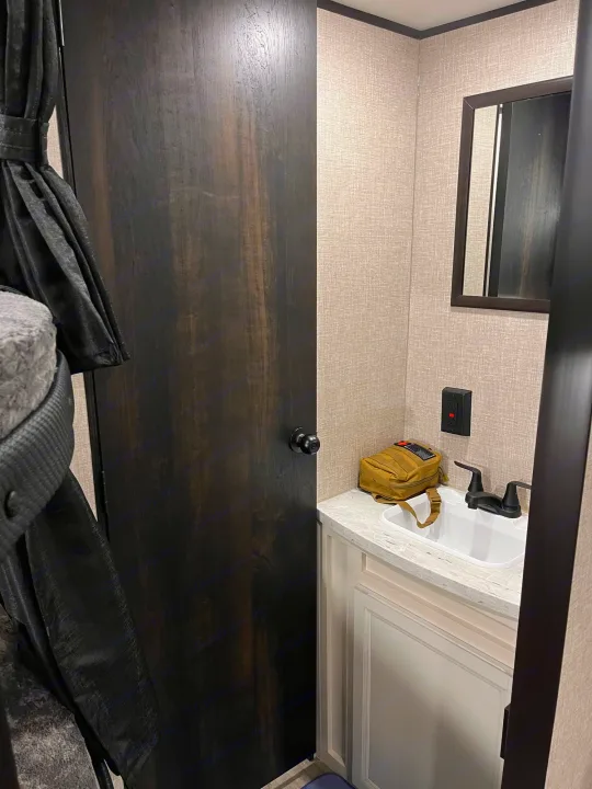 RV rental: Compact bathroom vanity with sink, mirror, and dark wood door. Bunk bed curtain visible left.. Jayco Jay Flight 2022