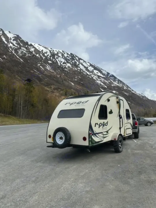 An r.pod RV rental travel trailer is parked with its spare tire visible, set against a stunning backdrop of snow-capped mountains.. Forest River R-Pod 2015