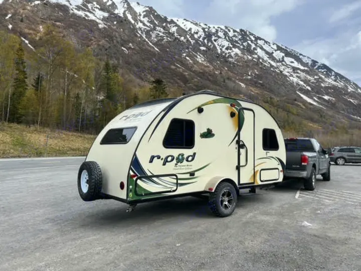 A cream-colored R-Pod RV rental travel trailer, hitched to a dark truck, is parked in front of snow-capped mountains and trees.. Forest River R-Pod 2015