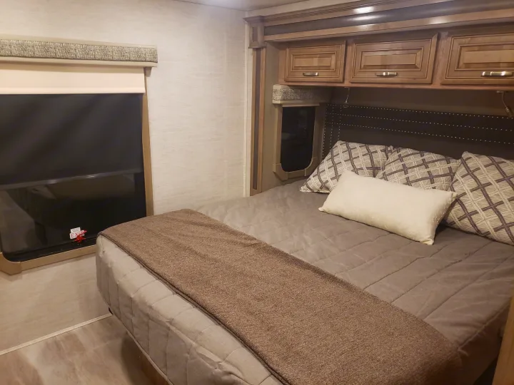 RV rental bedroom: cozy space with comfy bed, patterned pillows, overhead cabinets, and window with an exit handle.. Jayco Seneca 37L 2021