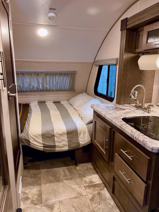 Cozy RV rental interior: a comfortable bed with striped sheets, compact kitchen with sink, and light-colored floor tiles.. R-Pod Hood River Edition 2017