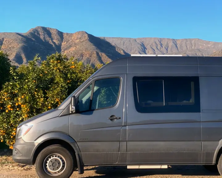 Gray RV rental van parked by a vibrant orange grove with scenic mountains in the background. Perfect for nature getaways.. Mercedes-Benz Sprinter 2016