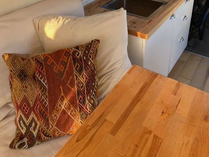 Cozy RV rental interior with patterned pillow, kitchenette, and wooden table, bathed in warm sunlight.. Mercedes-Benz Sprinter 2016
