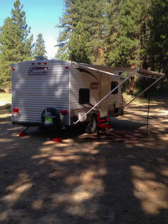 Shaded Coleman RV rental travel trailer at a serene campsite with an extended awning, mat, and chair, ready for relaxation.. Dutchmen Coleman Expedition LT 2013