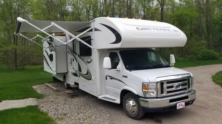 White Class C **RV rental** with awning extended and slide-out open, parked on gravel beside green grass and trees.. Jayco Greyhawk 2013