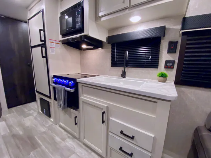 This compact RV rental kitchen features a modern fridge, microwave, stove with blue knobs, sink, and ample storage.. Jayco 264BH 2022