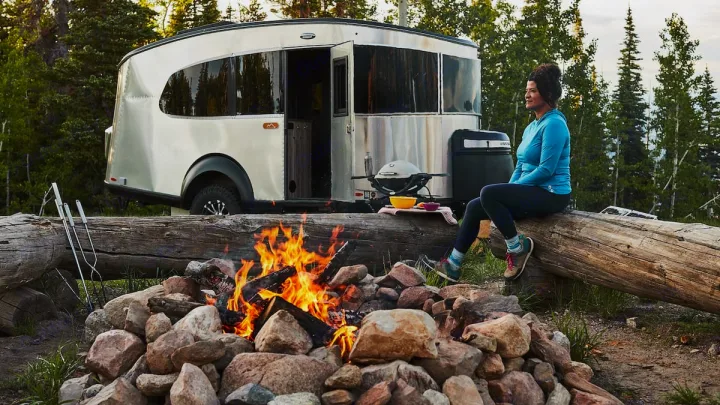 A woman relaxes by a campfire at an rv rental campsite, with a silver camper and grill in a forest.. Airstream Base Camp 2023
