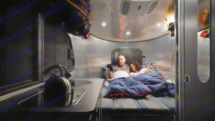 Cozy RV rental interior with a couple relaxing in bed; the man uses a tablet, kettle visible on the counter.. Airstream Base Camp 2023