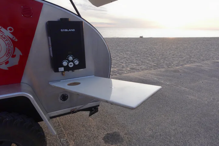 Visually, a red and silver teardrop **rv rental** is parked by a sandy beach, featuring an exterior water heater and a fold-out table.. Second Wind Freedom 2020