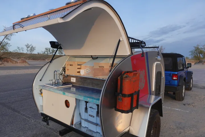 Discover this rv rental teardrop camper's kitchenette with sink, counter, and cooler. Parked by a Jeep in sandy terrain.. Second Wind Freedom 2020