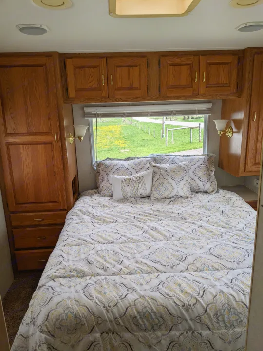 Cozy RV rental bedroom featuring patterned bedding, wooden built-in storage, and a scenic window view of a grassy field.. Holiday Rambler Atlantis 2003