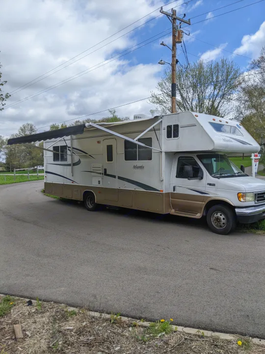White and tan Atlantis Class C RV with an extended awning, parked on an asphalt road. Ready for your next RV rental adventure.. Holiday Rambler Atlantis 2003
