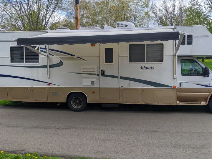 A white and gold Atlantis RV rental, with its awning extended, parked on asphalt with trees in the background.. Holiday Rambler Atlantis 2003