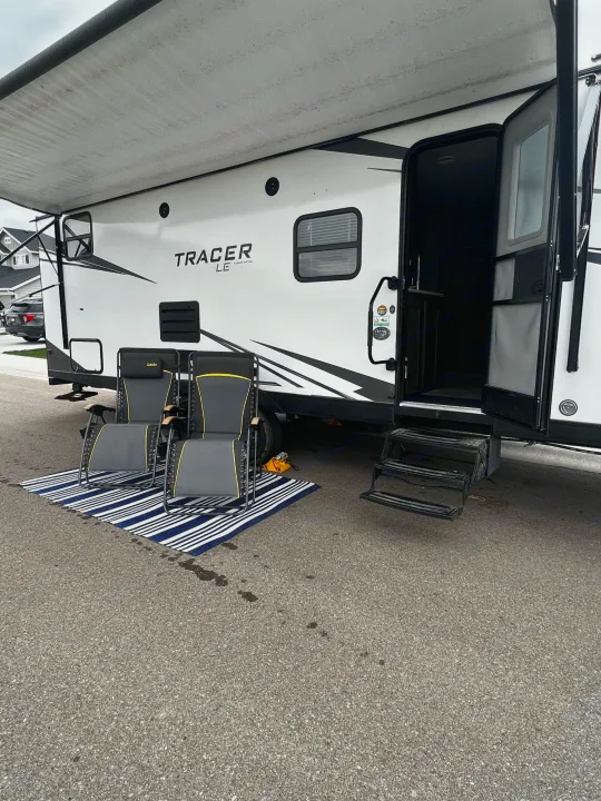Relaxing RV rental setup: white travel trailer with extended awning, two chairs on a mat, and open door.. Forest River Tracer LE 2020