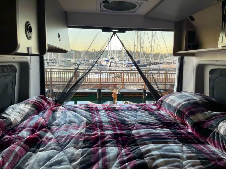 Relaxing in an RV rental, looking out at a stunning marina. Cozy plaid bed, boats, and docks create a serene waterfront view.. Winnebago Winnebago Solis 59P 2022
