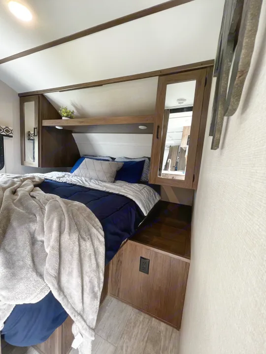 Cozy RV rental bedroom features a comfy bed with navy/grey bedding, overhead storage, and warm wood accents.. Forest River Tracer LE 2020