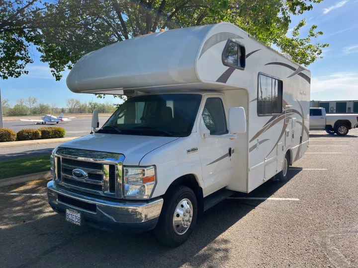 White Class C RV parked in a sunny lot, small airplane on a runway in the distance. Ideal for your next rv rental.. Thor Four Winds 2013