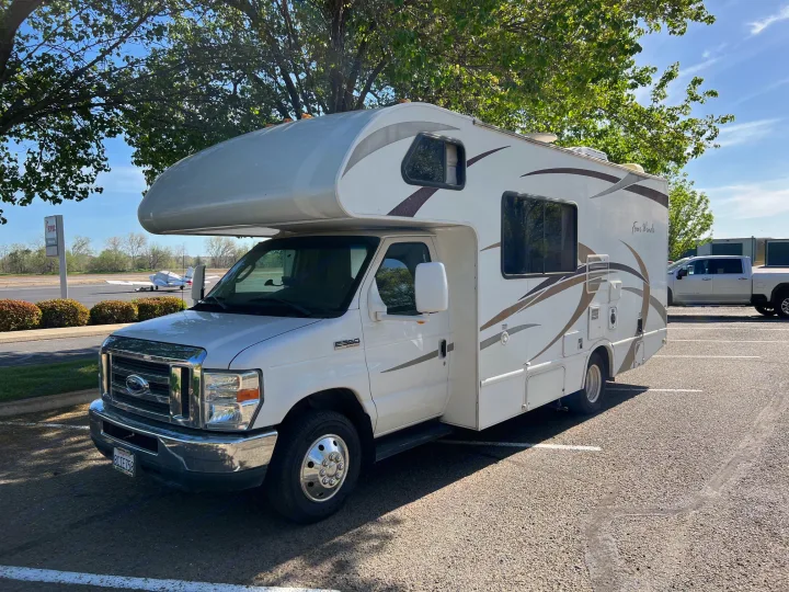 White RV rental (Ford Class C motorhome) parked in a lot. An airplane is visible on a tarmac in the background.. Thor Four Winds 2013