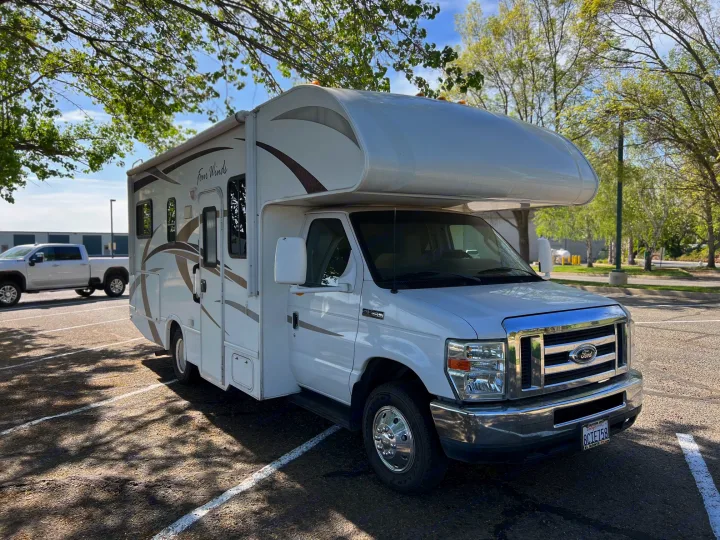 White Ford Four Winds Class C rv rental parked in a sunny lot, shadows from overhead trees on the pavement.. Thor Four Winds 2013