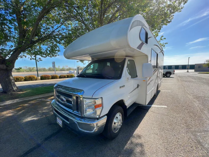 A pristine white Ford E-350 Class C RV rental, a motorhome, parked under sunlit trees in a spacious lot.. Thor Four Winds 2013