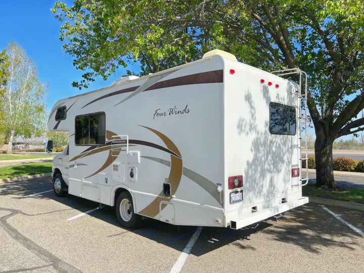 This white "Four Winds" RV rental is parked under a large tree on a sunny day, showcasing its side and back.. Thor Four Winds 2013