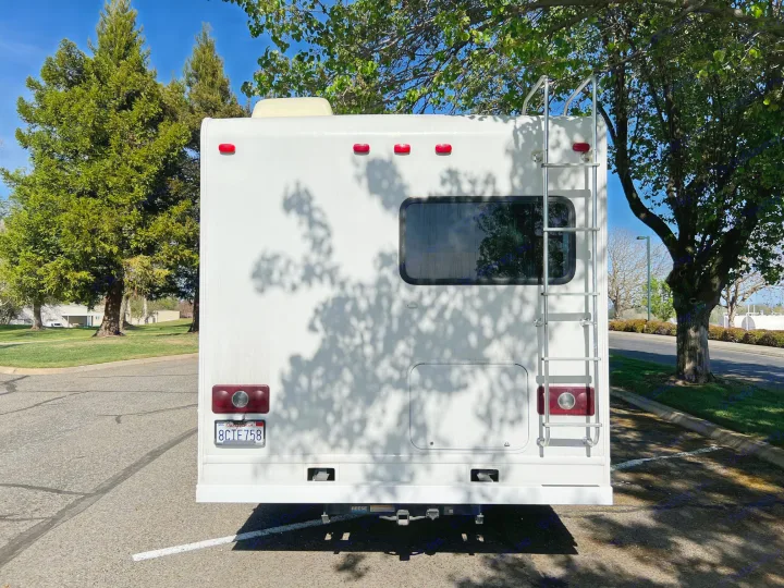 Back of a white RV rental, showing ladder, tail lights, and CA license plate (8CIE758) in a sunny parking lot.. Thor Four Winds 2013