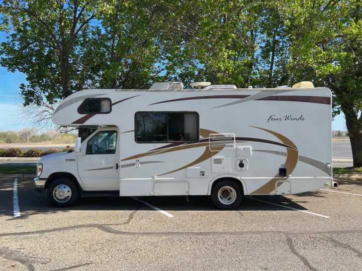 White Class C RV rental, a Four Winds motorhome, parked on asphalt with green trees and blue sky in the background.. Thor Four Winds 2013