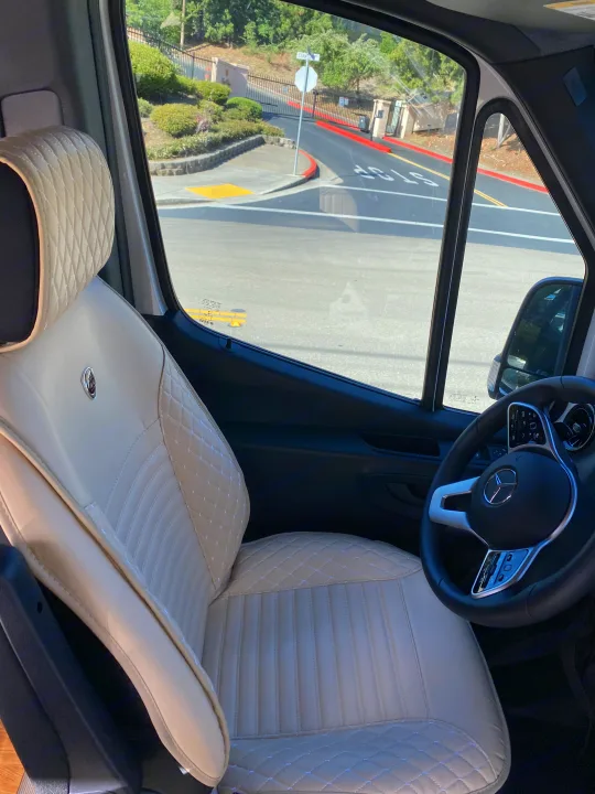 Inside an RV rental: luxurious cream quilted driver's seat and Mercedes-Benz steering wheel, with street view visible.. Vanworks Mercedes Benz Sprinter 2021