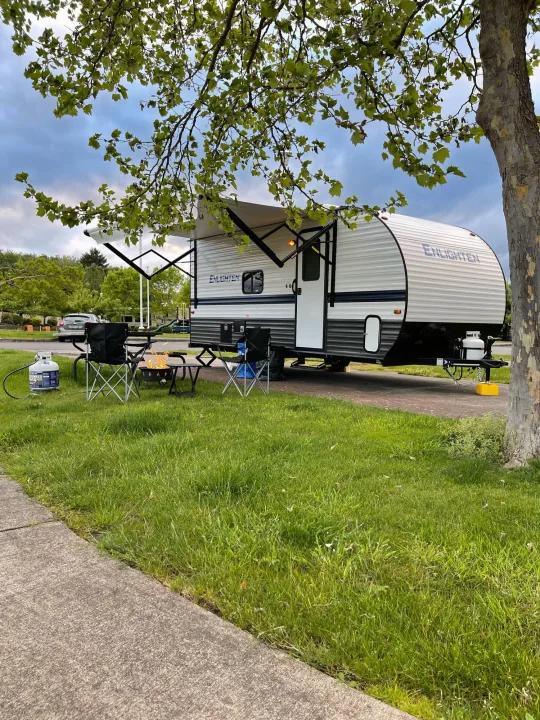 An RV rental parked on a paved site, with camping chairs and a lit propane fire pit on lush green grass. Shady trees overhead.. Gulf Stream Enlighten 18BH 2022