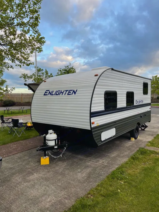An "Enlighten" travel trailer RV rental is set up on a concrete pad for camping, complete with chairs and a portable fire pit.. Gulf Stream Enlighten 18BH 2022