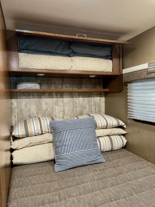 RV rental cozy bedroom: overhead storage with bedding, open shelves, and a bed with pillows for compact living.. Winnebago Outlook 2019