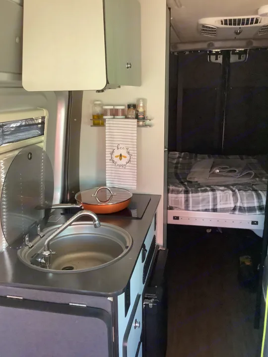RV rental interior: Compact camper van kitchen with sink, cooktop, and cozy sleeping area beyond.. Winnebago Revel 2019