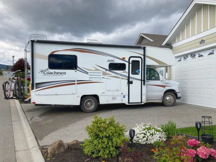 White Class C RV rental with bikes on the back, parked on a residential driveway next to a house under a cloudy sky.. Coachman Freelander 2013